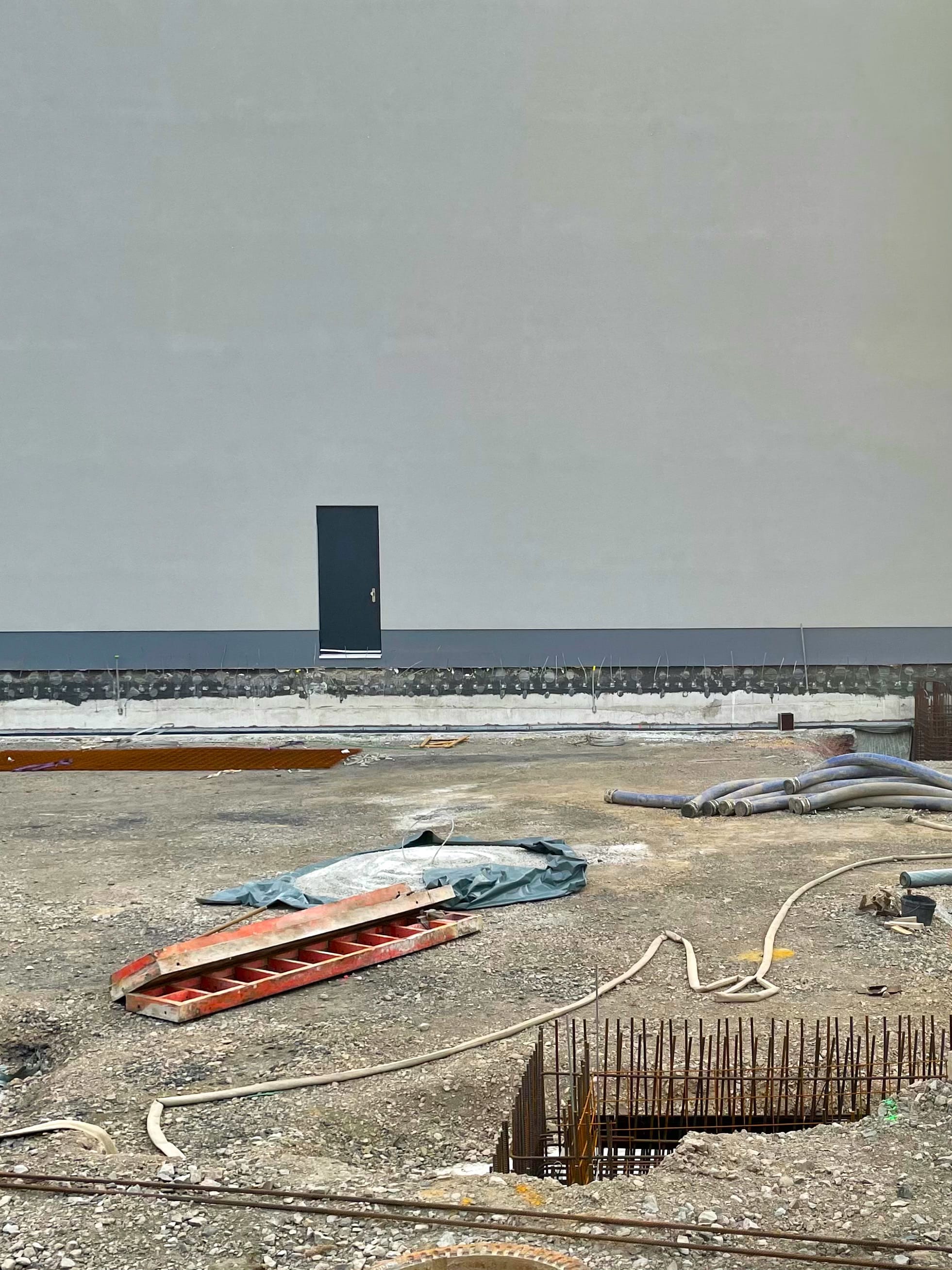 Baustelle Dortmund, eine scheinbar kleine Tür an einer hellgrauen Wand, im Vordergrund mehrere Rohre, ein sich windender Schlauch und Stahlrohrgitter sowie ein rotes kassetiertes Holzelement, platziert wie Kunstwerke.