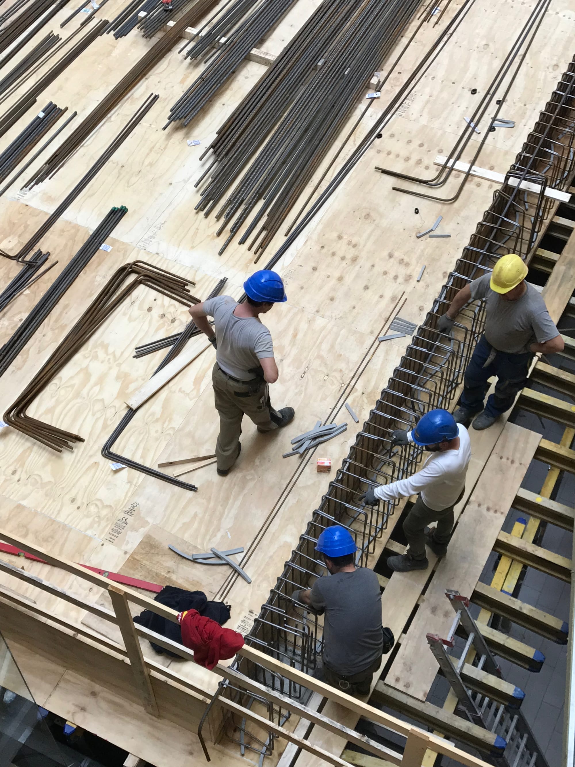 Baustelle von oben, vier Bauarbeiter mit Helmen installieren auf einem Holzaufbau Metallstreben, eine Leiter führt nach unten, ein roter Pullover liegt über einem Holzgeländer